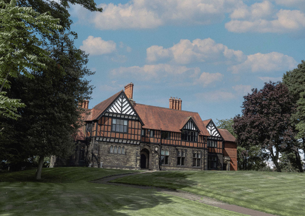 A Tudor-style manor house sits atop a gentle, grassy slope, framed by mature trees and shaded lawns, beneath a blue sky scattered with soft, white clouds.