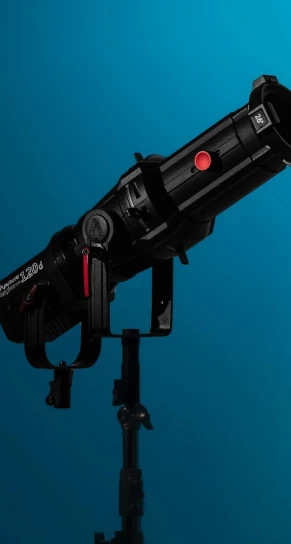 Studio spotlight mounted on an adjustable stand, angled diagonally upward; black barrel has a red circular sticker and focusing yoke against a smooth teal-blue gradient background. Text visible: "Aputure 120d" and "75".