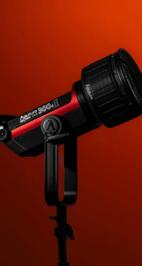 Studio LED light (Aputure 300d II) mounted on a stand, tilted slightly and aimed right against a deep red gradient background. Text visible: "Aputure 300d II" and "Aputure".