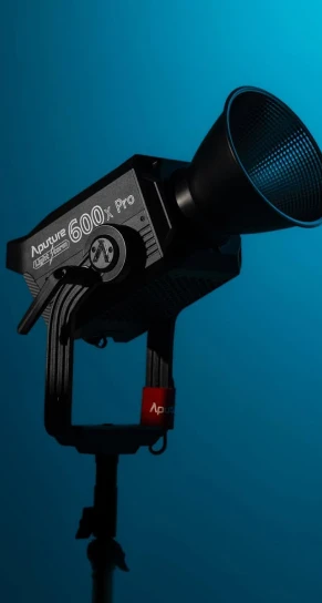 Aputure 600x Pro studio light sits angled on a yoke-mounted stand with a conical reflector attached, pointing slightly downward against a teal gradient studio background. Transcribed text: "Aputure 600x Pro", "Aputure"